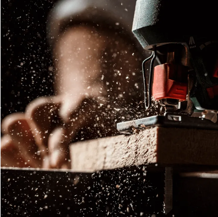 Fabrication sur mesure en atelier de menuiserie : gros plan d'une scie sauteuse découpant une planche de bois, projection de sciure dans l'atelier sombre.