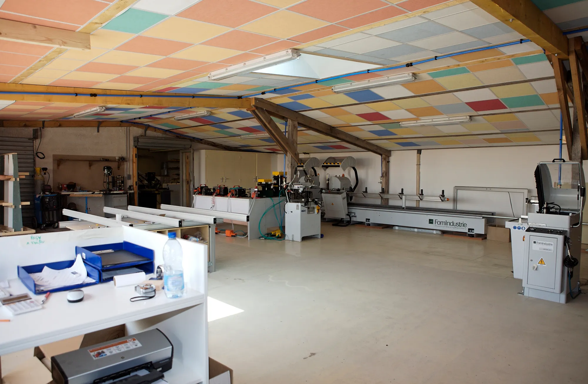 Fabrication sur mesure en atelier de menuiserie : plafond en panneaux colorés, plaqueuse de chants et longs établis sous charpente en bois