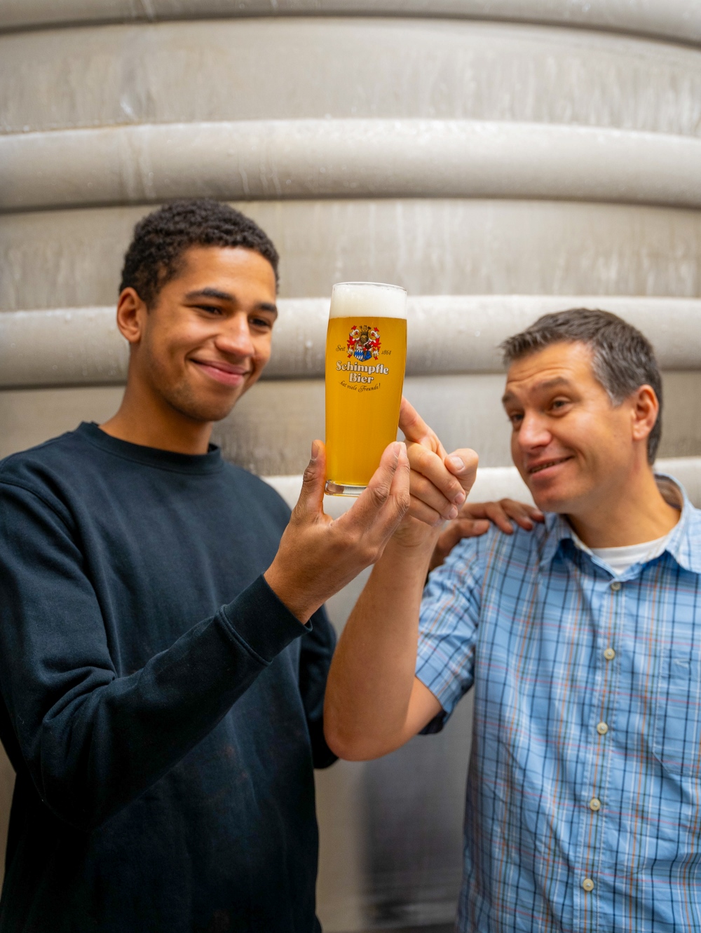 Zwei Männer stehen nebeneinander und halten ein Glas Bier. Der Mann rechts zeigt darauf.