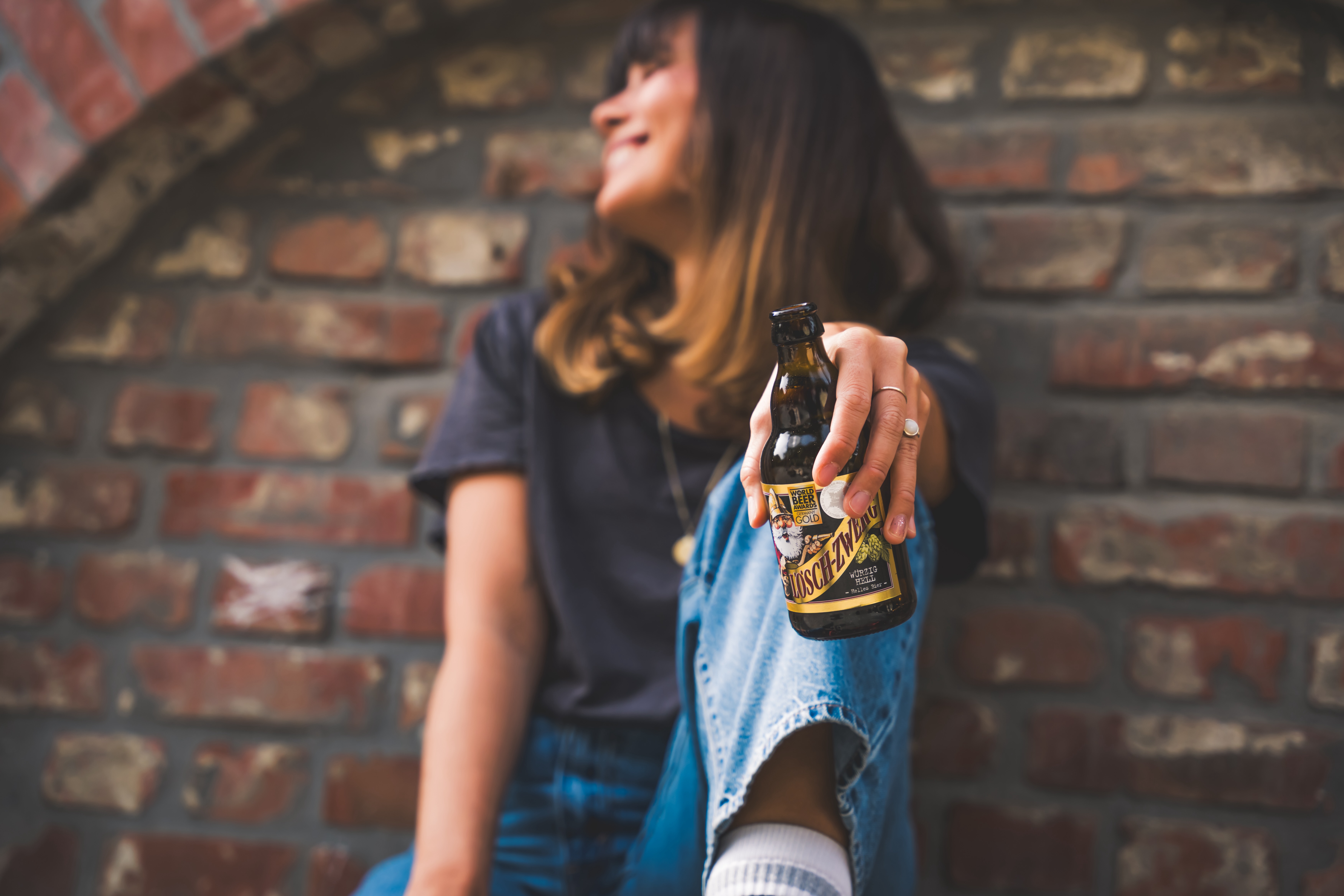 Eine Frau sitzt angelehnt an einer Wand und hält lässig eine Lösch-Zwerg-Flasche.