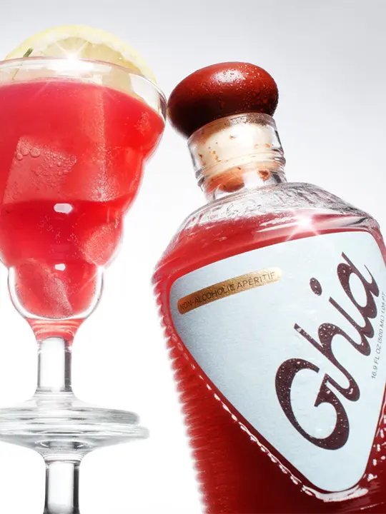 Red Ghia non-alcoholic aperitif bottle next to a red cocktail glass garnished with a lemon slice.