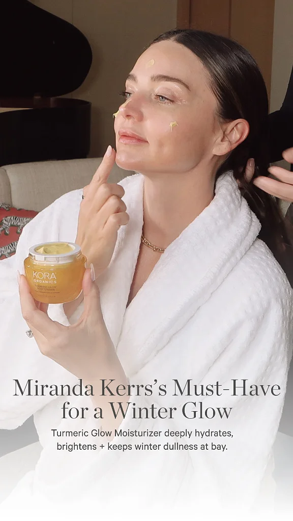 Woman in white robe applying Kora Organics Turmeric Glow moisturizer to her face with dots on cheeks and forehead.