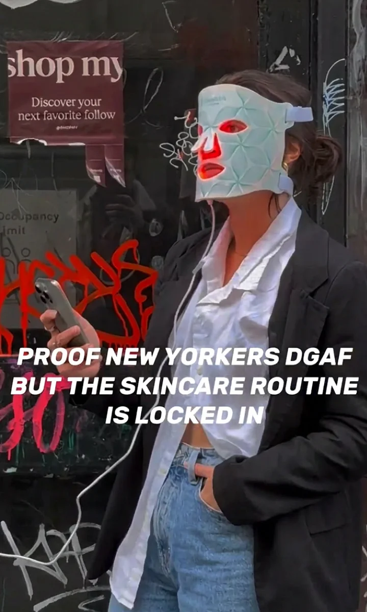 Person wearing a white LED facial mask and holding a phone in front of a graffiti-covered wall.
