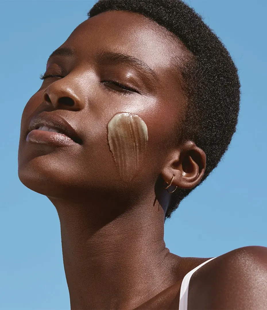 Portrait of a woman with closed eyes and skincare cream swiped on her cheek against a blue sky.