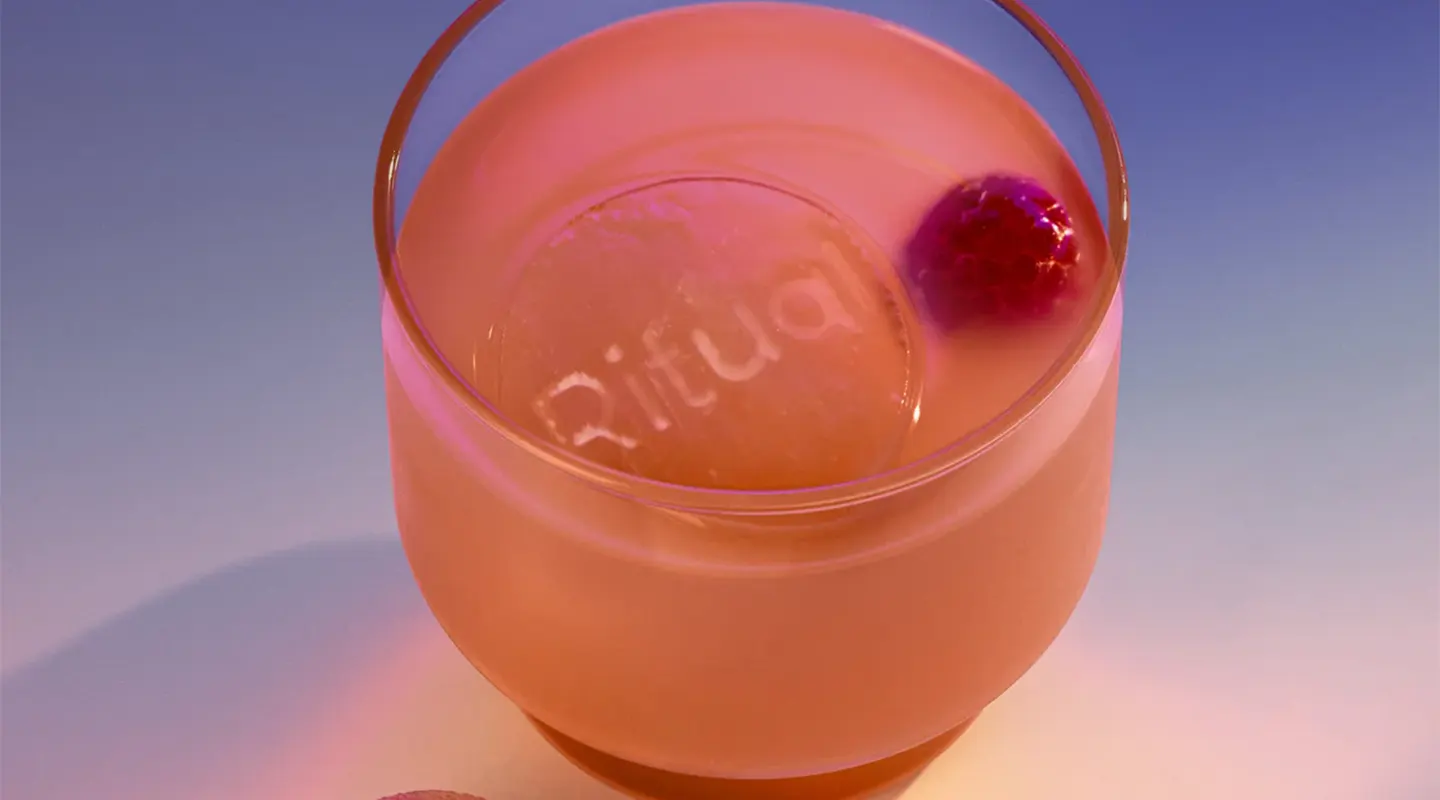 A pink cocktail with a large ice cube labeled Ritual and a raspberry in a clear glass.