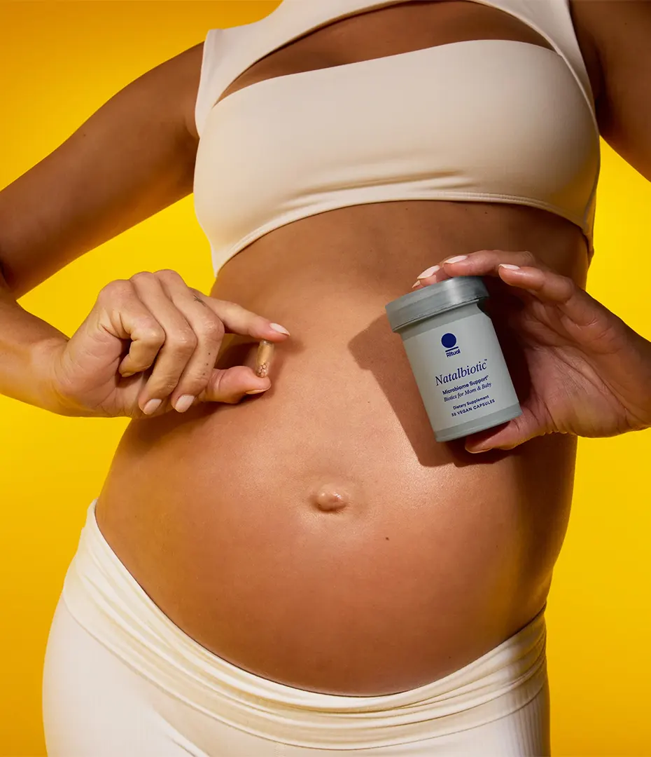 Pregnant woman in beige outfit holding a Natalbiotic supplement bottle and capsule near her belly.