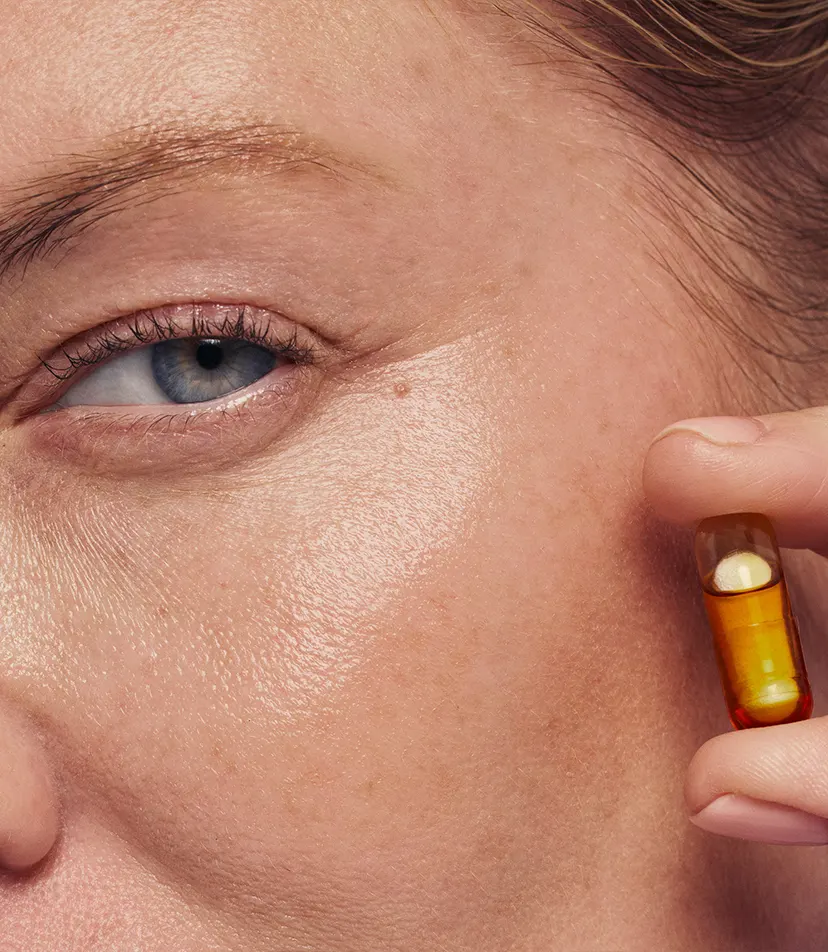 Close-up of a person holding a yellow and amber capsule near their cheek and eye.