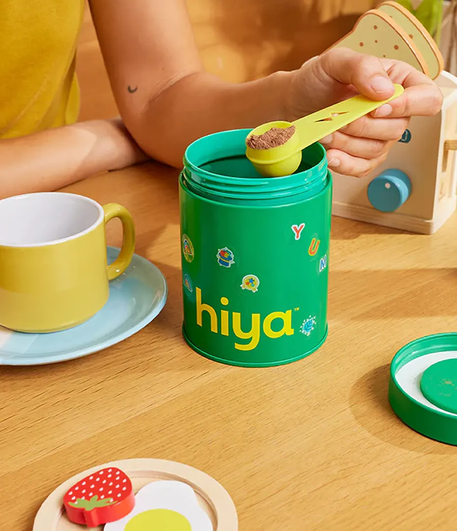 Hand holding yellow scoop of brown powder over a green container labeled hiya on a wooden table.