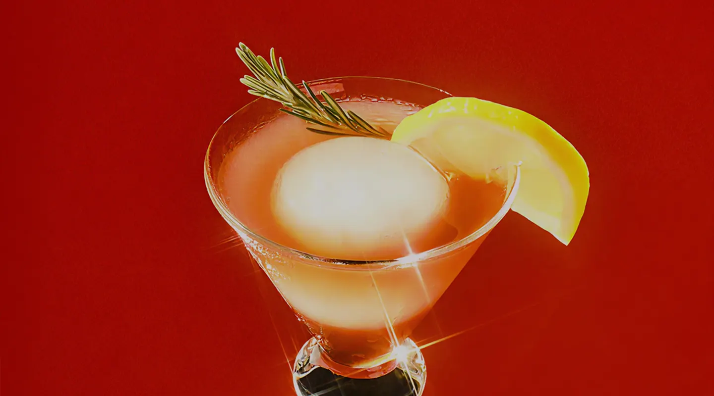 Martini glass with an orange cocktail, a large round ice ball, a lemon wedge, and rosemary garnish.