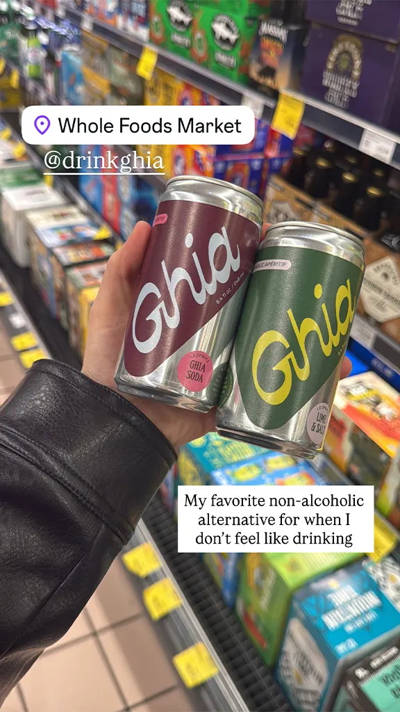 Hand holding two cans of Ghia non-alcoholic soda in a Whole Foods Market aisle.