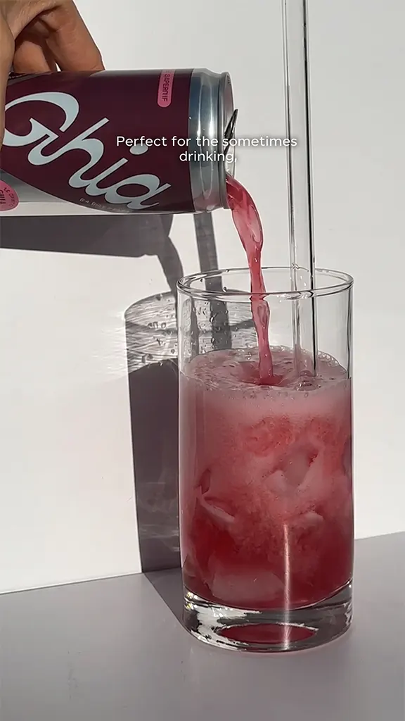 Hand pouring a pink Ghia drink from a can into a glass with ice on a white surface.