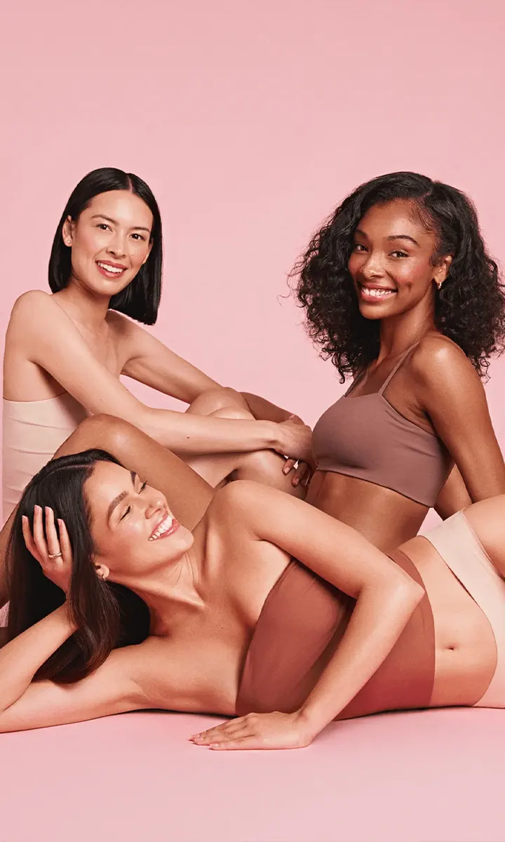 Three diverse women in neutral-toned underwear posing and smiling against a pink background.