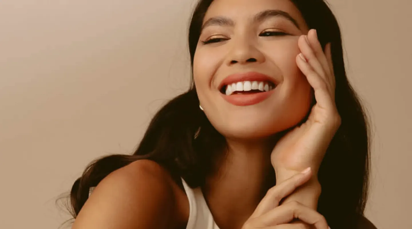 Smiling woman with long dark hair touching her face against a beige background.