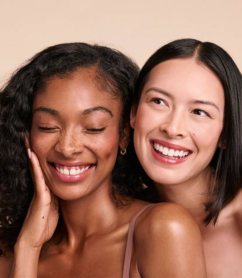 Two smiling women with clear skin, one with curly hair and freckles, the other with straight hair.