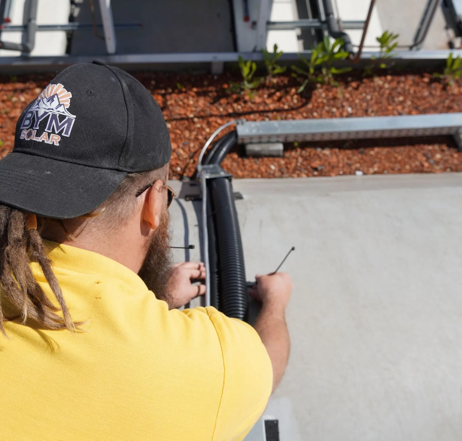Techniker in gelbem T-Shirt montiert Solarpanels auf einem Dach vor grünem Laub im Sonnenschein.