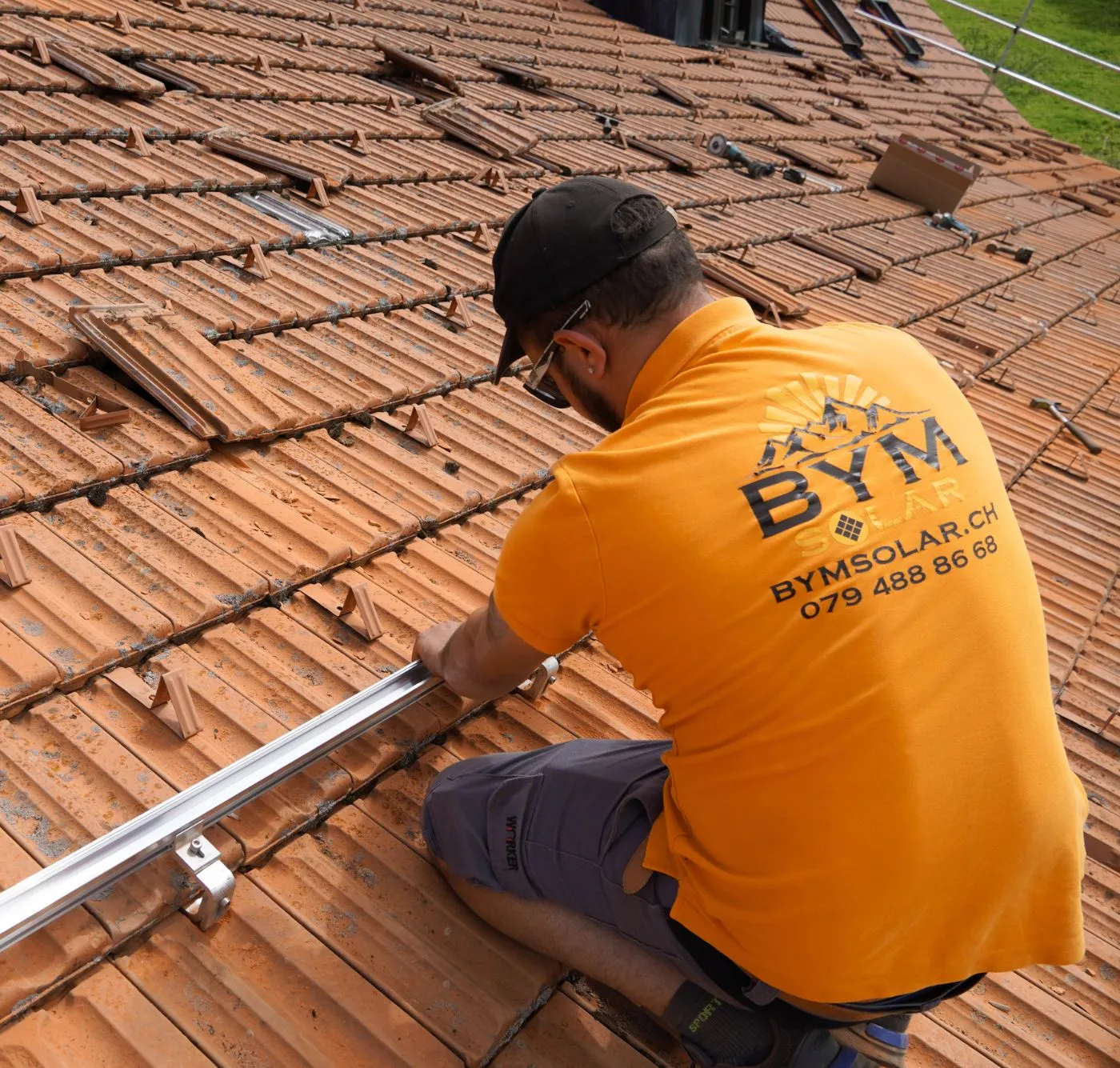 Ein Handwerker in orangefarbenem BYM Solar Poloshirt montiert eine Aluminiumhalterung auf einem Ziegeldach.