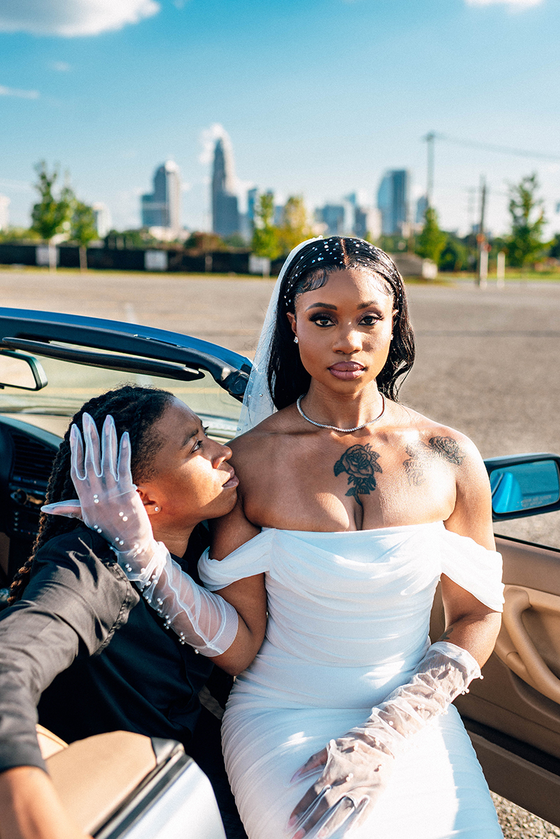 Elopement styled shoot in Camp North End Charlotte, NC with same-sex couple in classic BMW convertible. Couple sitting in passenger's seat, bride sitting on other partner's lap. Bride looking at camera