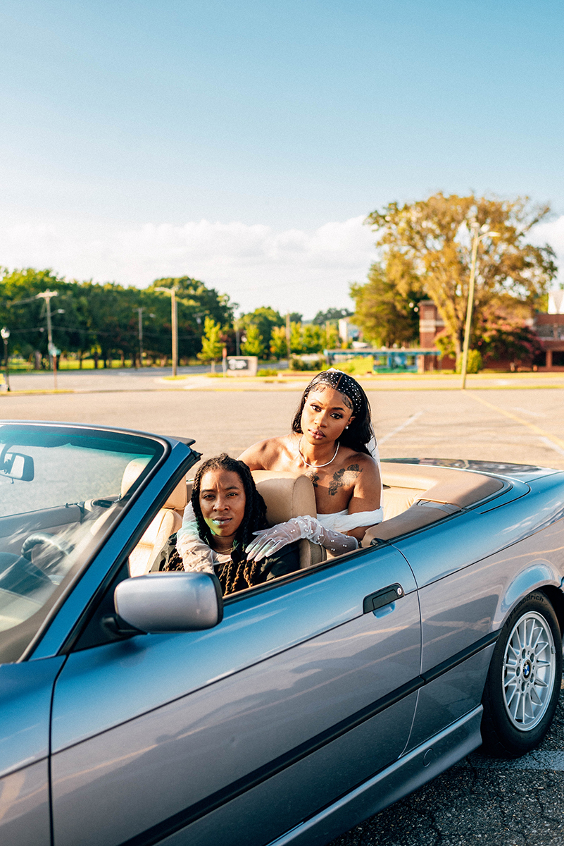 Elopement styled shoot in Camp North End Charlotte, NC with same-sex couple in classic BMW convertible. Bride in backseat leaning over other partner in driver's seat