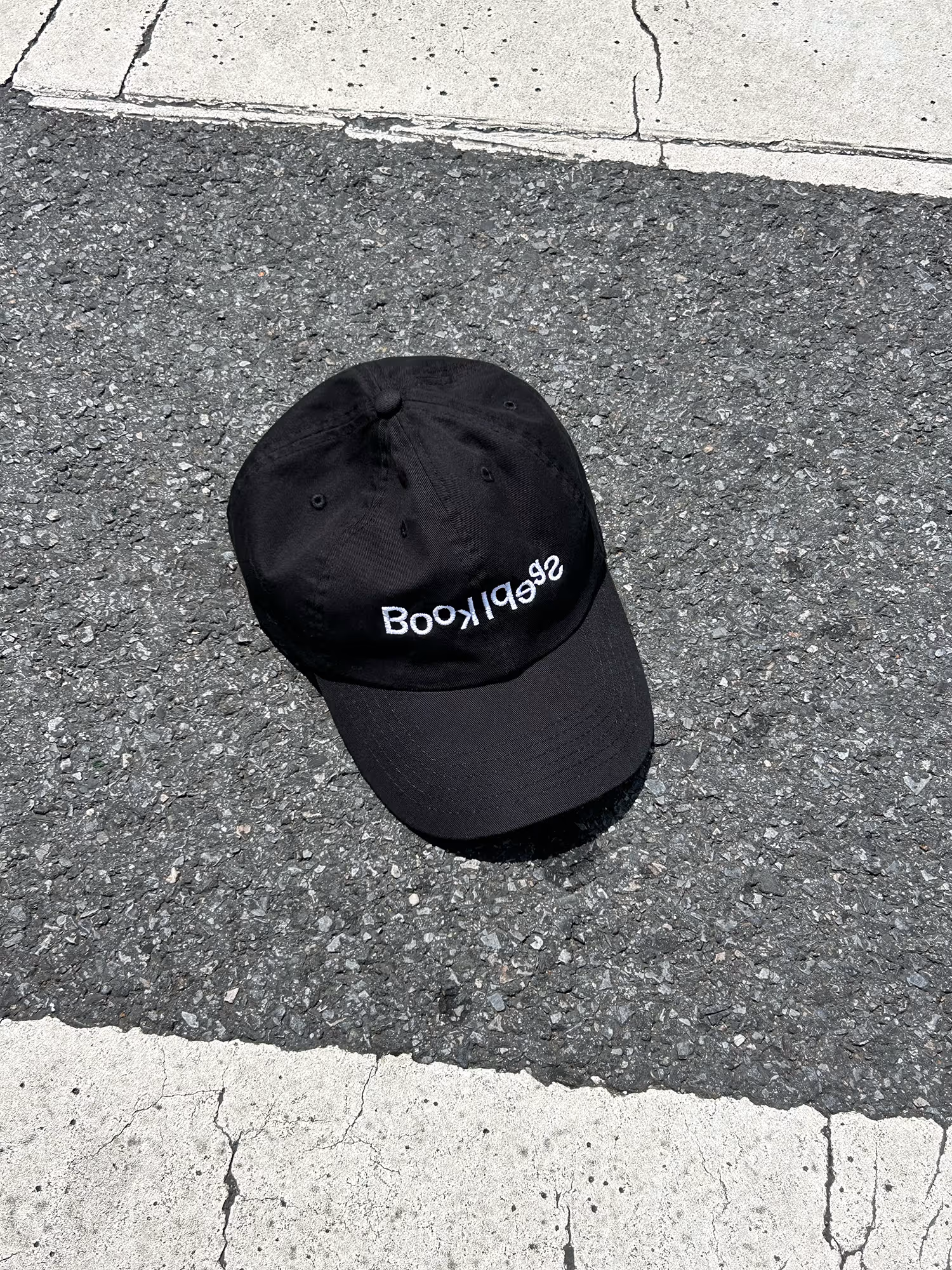 A black baseball cap with the word "Book Ideas" embroidered in white, lying on an asphalt road partially within a white road marking.