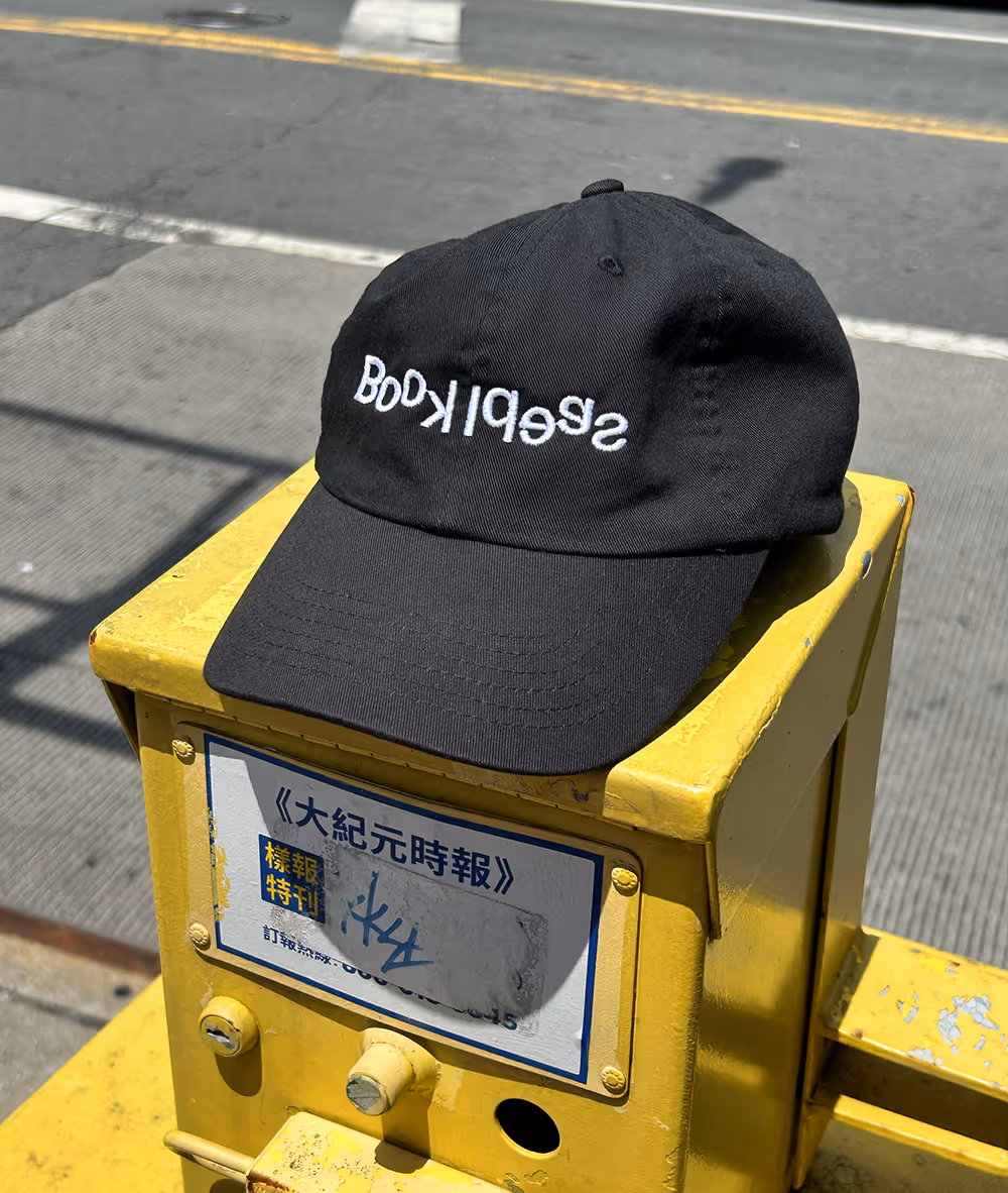 A black baseball cap with the word "Book Ideas" embroidered in white, resting on top of a bright yellow newspaper vending box on a city sidewalk.