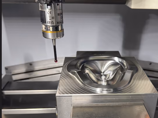 Probe on a coordinate measuring machine inspecting a shiny metal mold die with curved contours.