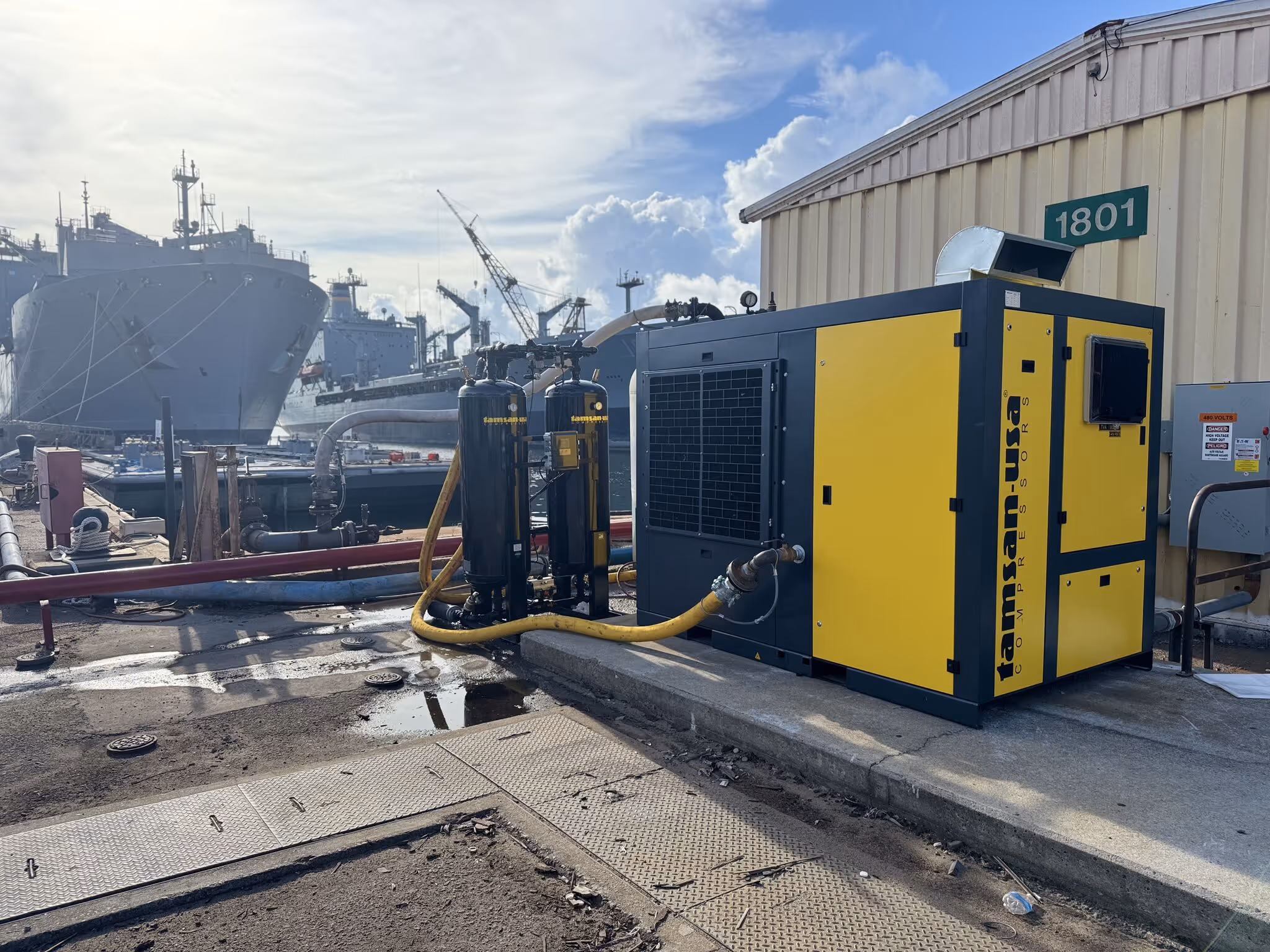Tamsan USA industrial air compressor is operating outdoors near a dock, connected with yellow hoses to two vertical filtration units. In the background, several large naval ships and cranes are visible, along with a beige building marked “1801.