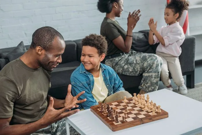 An image of a father teaching his son to play chess while family members celebrate in the background.