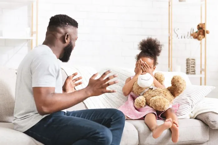 A view of a father scolding his young daughter as she covers her face with a teddy bear on the couch.