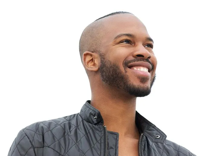 An image of a smiling young man in a black quilted jacket looking upward against a clear sky.