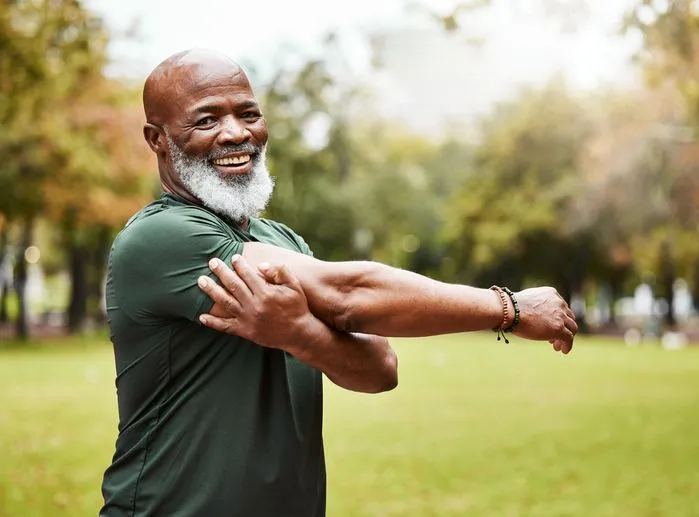 An image of a smiling older man with a white beard stretching his arm outdoors in a park.