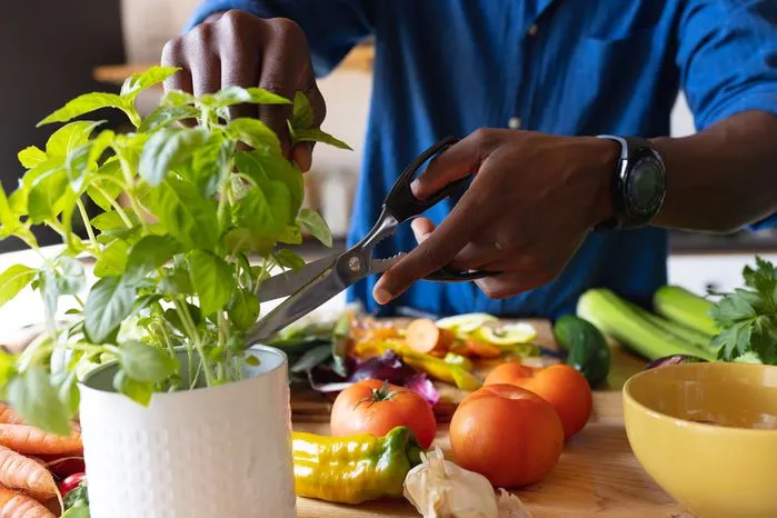 An image of a person cutting fresh basil leaves from a pot surrounded by colorful vegetables on a kitchen counter.