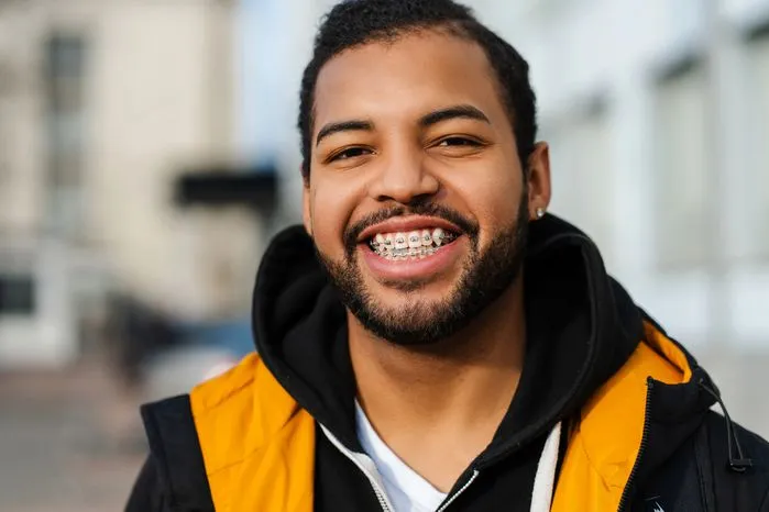 An image of a smiling young man with braces wearing a yellow jacket outdoors.