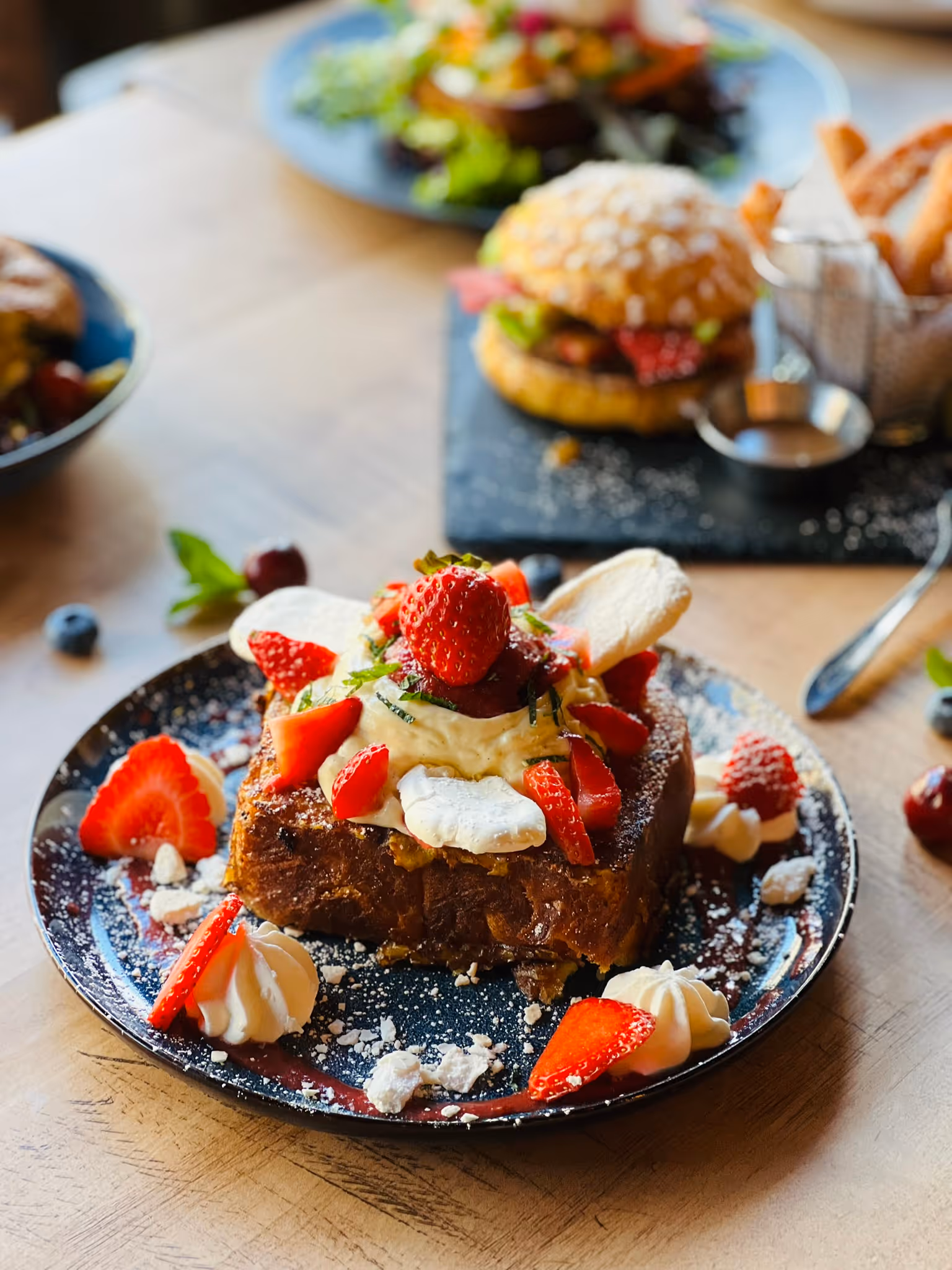 Pain perdu garni de fraises fraîches, crème fouettée et morceaux de meringue sur une assiette noire, avec un hamburger sucré et des frites de churros en arrière-plan flou.