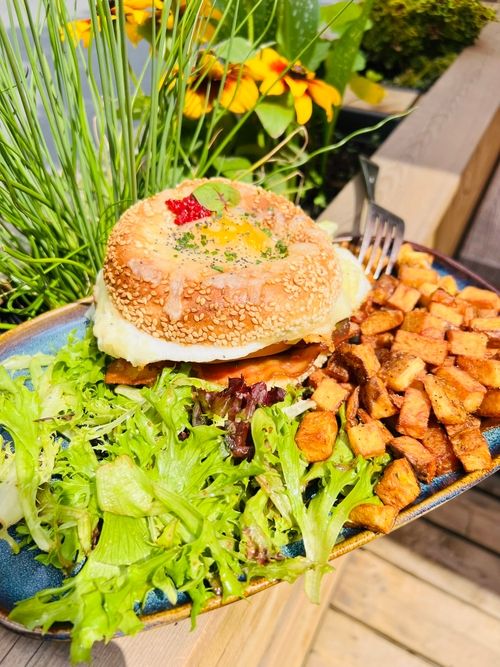 Bagel au sésame avec œuf, bacon, salade verte et pommes de terre rôties sur assiette bleue.