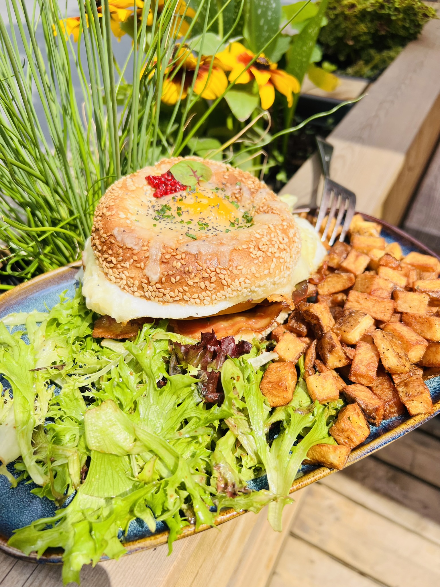 Bagel au sésame avec œuf, bacon, salade verte et pommes de terre rôties sur assiette bleue.