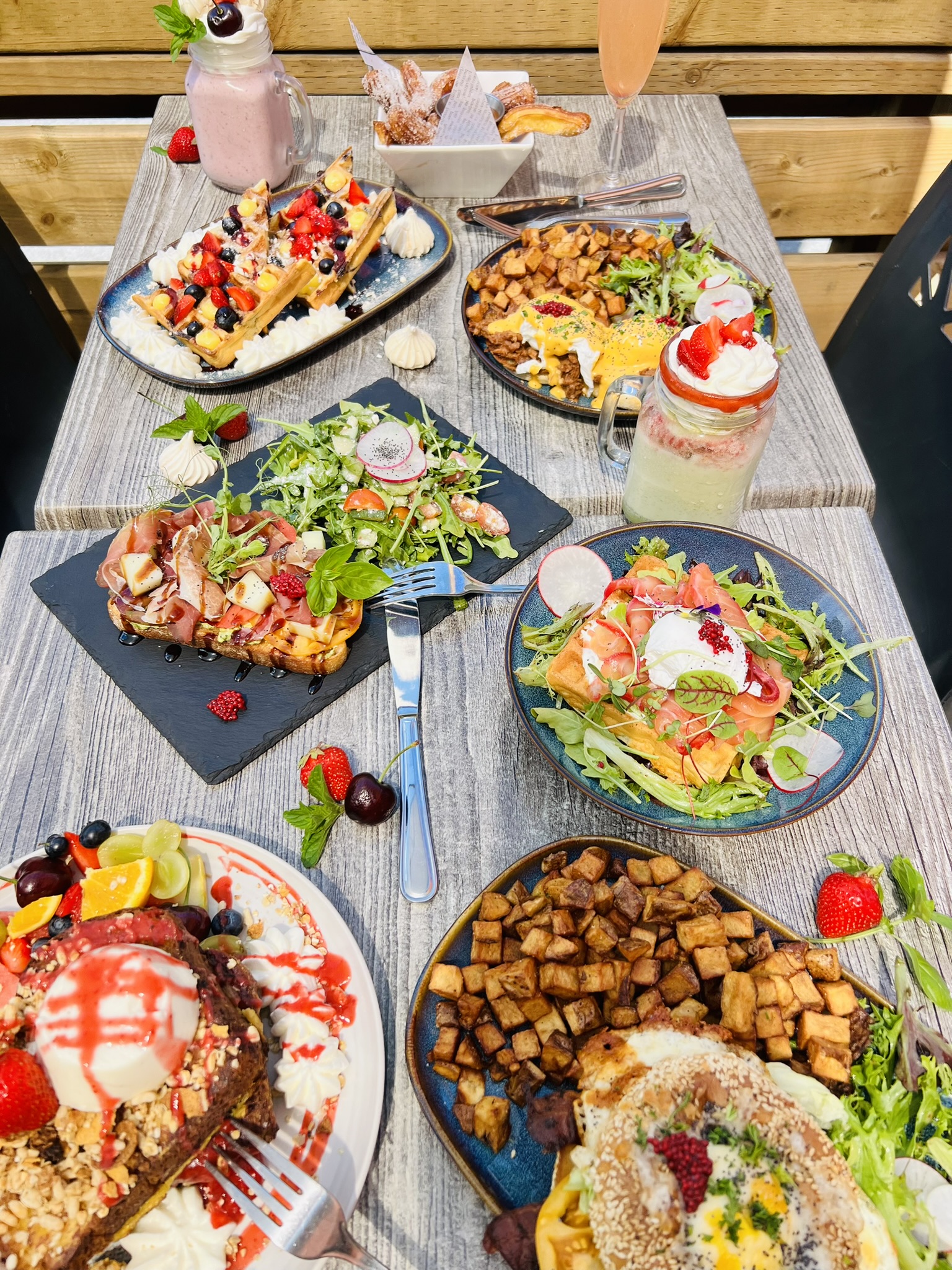 Table with breakfast dishes including waffles, eggs benedict, salad, potatoes, and fruit smoothies.