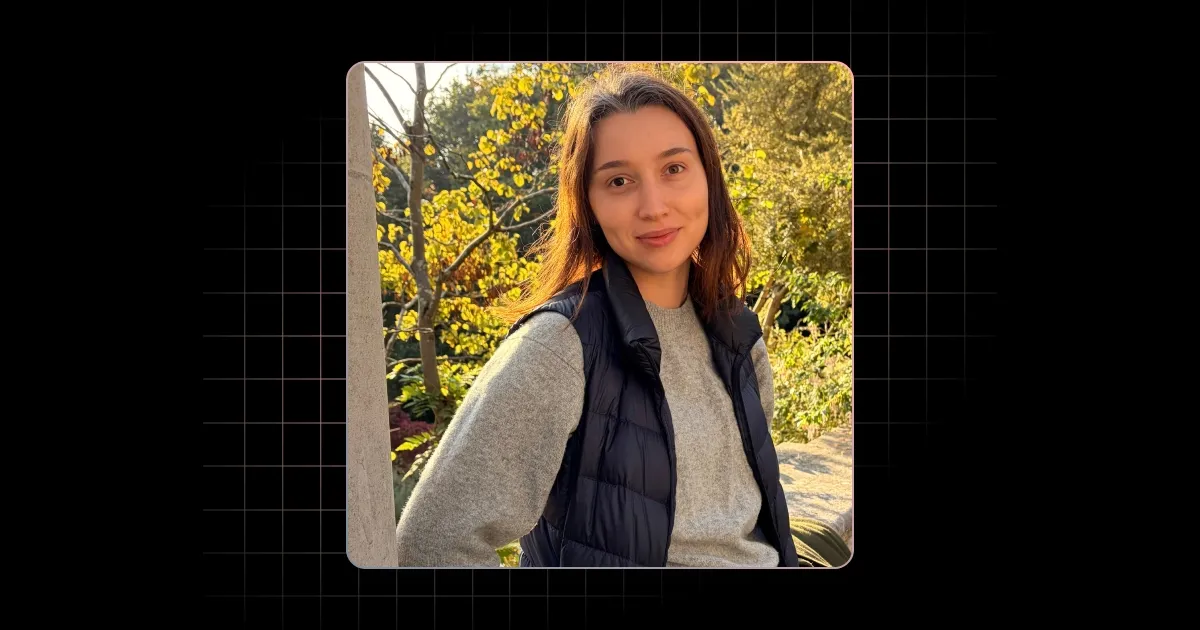 Young woman outdoors wearing a gray sweater and black puffer vest with trees in background.