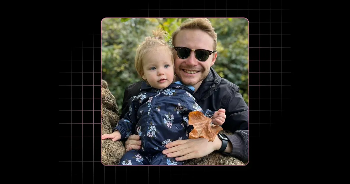 Smiling man wearing sunglasses holds a toddler dressed in floral clothes holding a dried leaf outdoors.