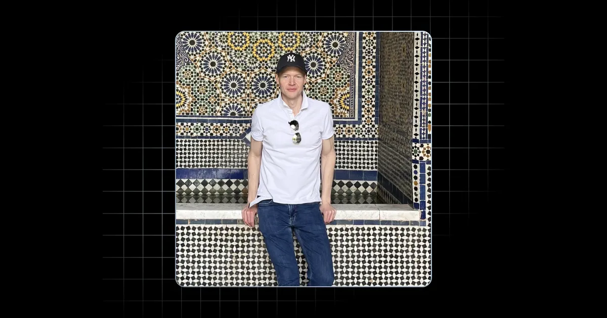 Man in white shirt and blue jeans sits in front of detailed mosaic tile wall wearing a black cap.