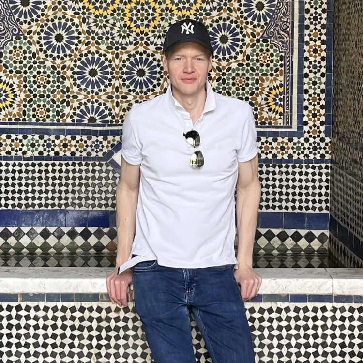 Man in white shirt and blue jeans wearing a black cap, standing against a colorful mosaic tile wall.