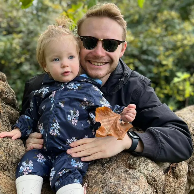 Smiling man wearing sunglasses holds a toddler dressed in floral clothes sitting on a tree branch.