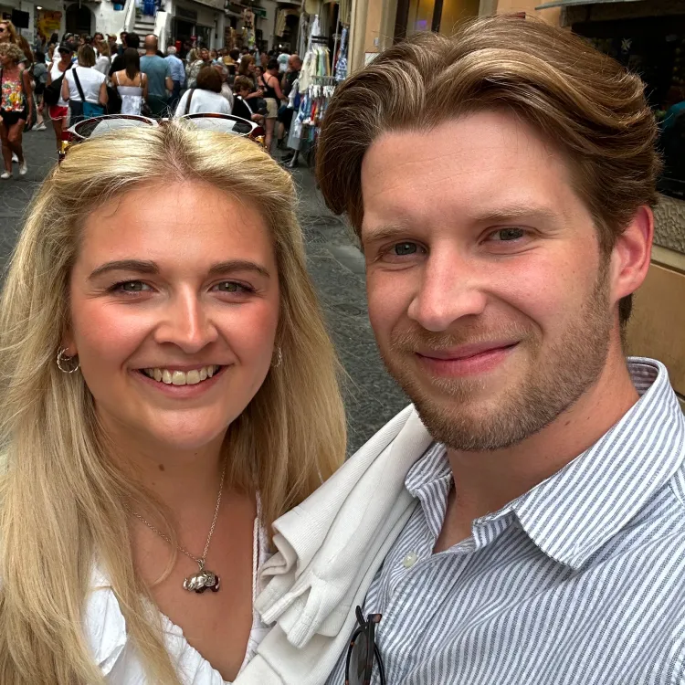 Smiling young man and woman take a selfie on a busy narrow street with people walking behind them.