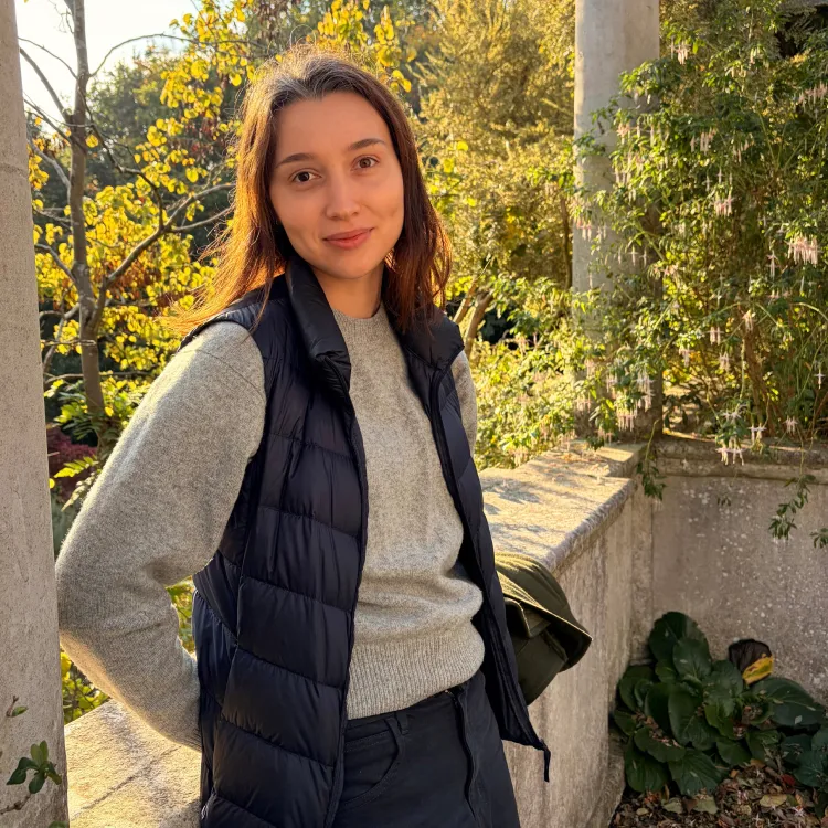 Young woman in a gray sweater and black vest standing outdoors with autumn foliage behind her.