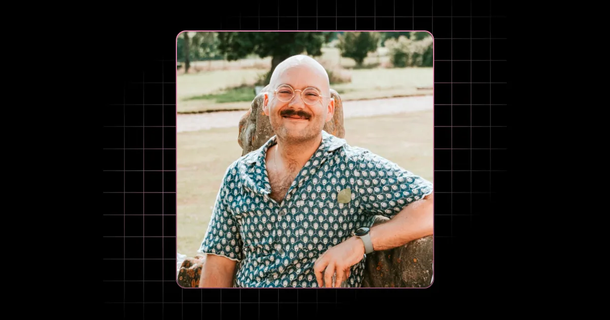Smiling bald man with mustache and glasses wearing a patterned shirt sitting outdoors on a stone bench.