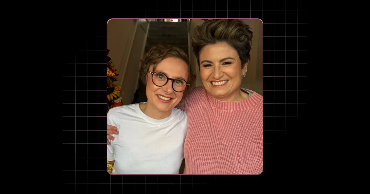 Two smiling women posing indoors; one in a white shirt and glasses, the other in a pink sweater.
