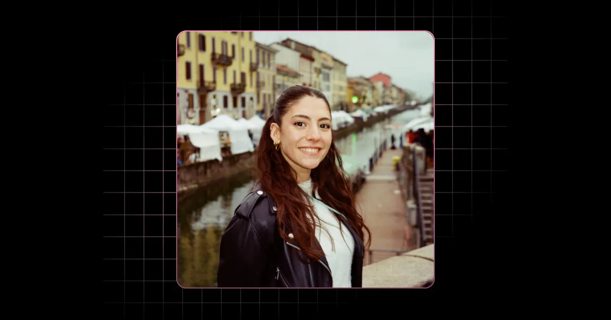 Woman with long hair and leather jacket smiling by a canal with buildings and tents in the background.