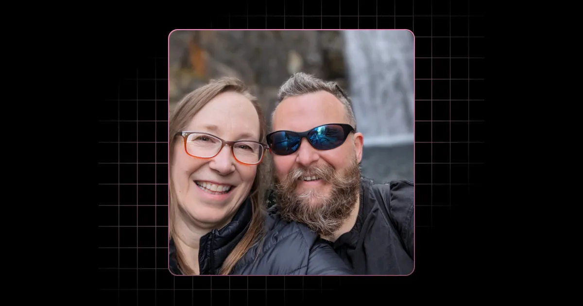 Smiling man with beard and sunglasses and woman with glasses wearing jackets near a waterfall.