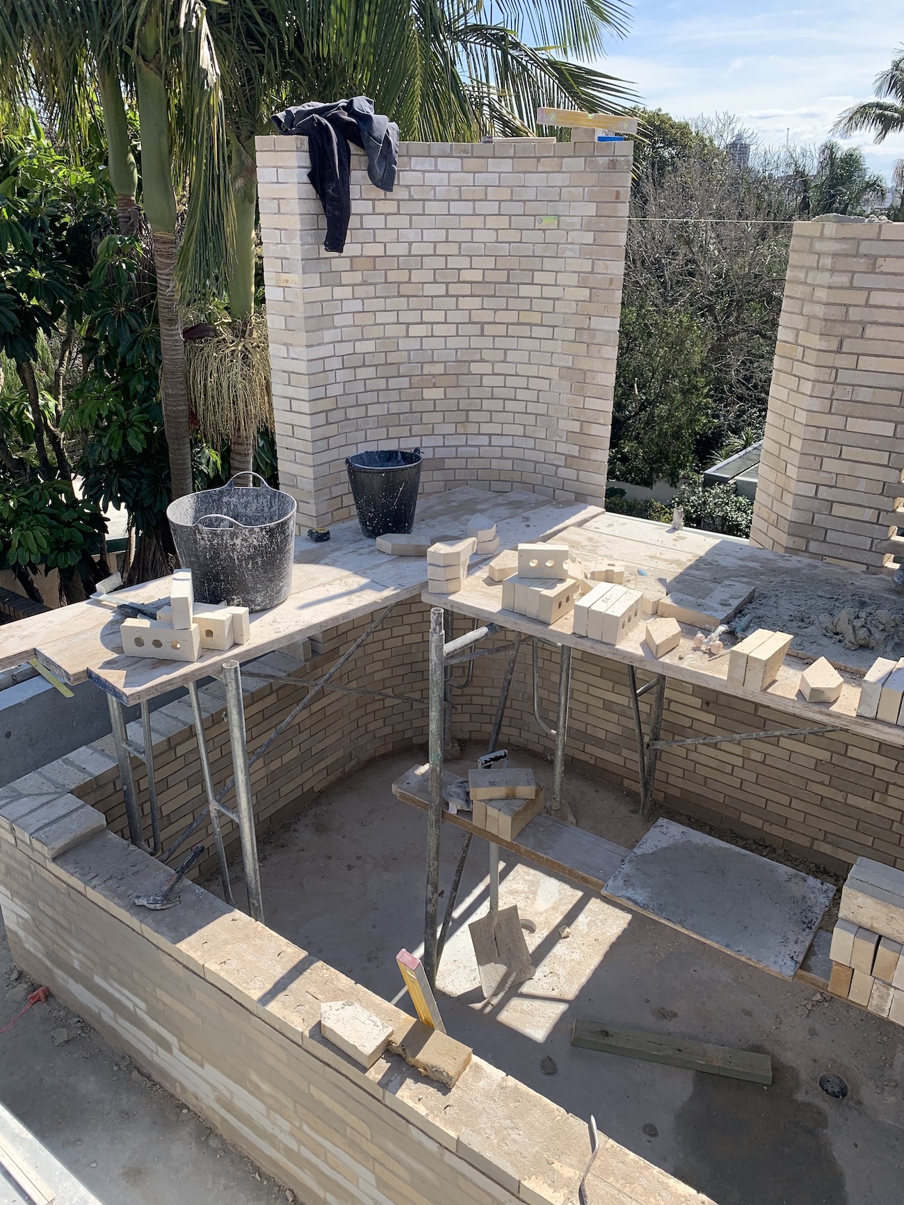 Construction site showing partially built brick structure with scaffolding, bricks, buckets, and a shirt hanging on the wall under palm trees.