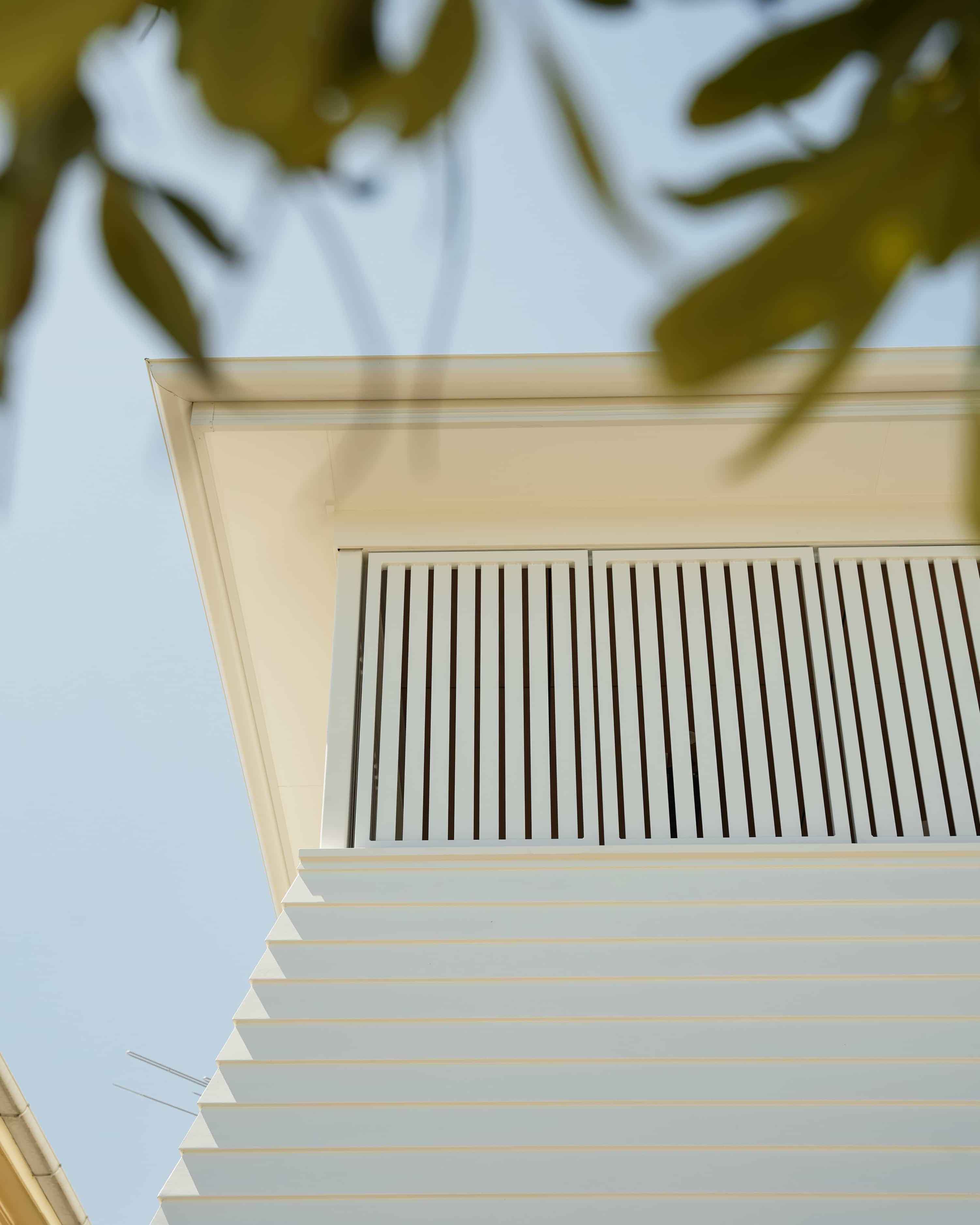 Bottom-up view of a white modern building facade with horizontal panels and vertical slats under a clear blue sky with blurred leaves in the foreground.
