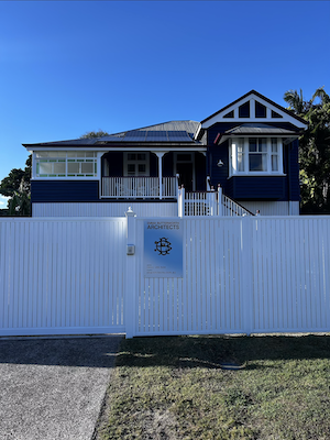 Two-story blue house with white fence and sign reading 'Architects' on the gate under a clear blue sky.